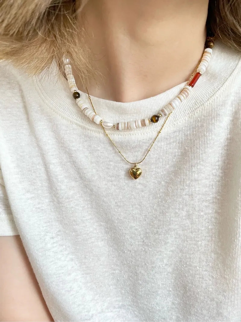 Woman in a white tee layering our natural shell bead necklace with a delicate gold heart pendant for a casual, everyday look.