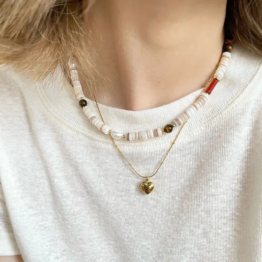 Woman in a white tee layering our natural shell bead necklace with a delicate gold heart pendant for a casual, everyday look.